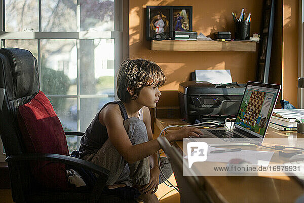 A preteen boy sits at computer playing an online chess game at desk