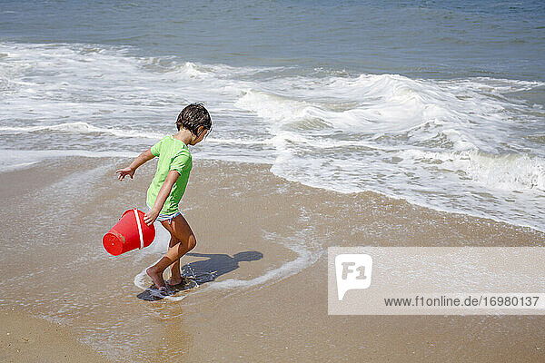 A little girl with a bucket plays in the tide at edge of sandy beach