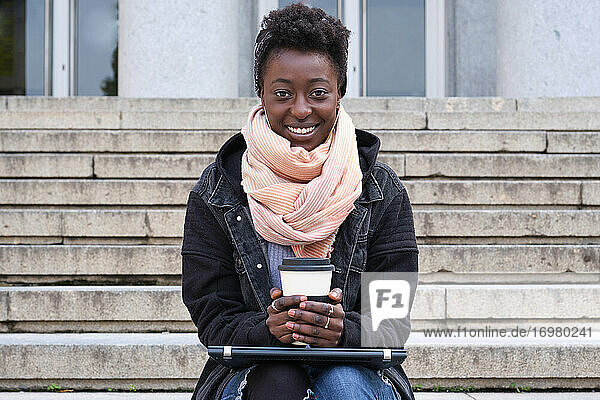 Schöne Universität afrikanische Studentin lächelnd Blick auf die Kamera  hält eine Tasse Kaffee  sitzen auf der Treppe außerhalb auf dem Campus. College Leben Konzept.