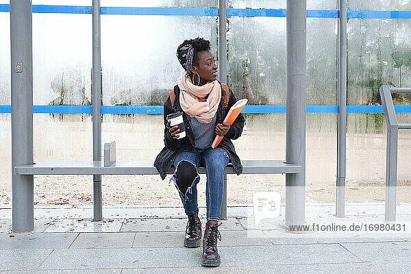 Universität afrikanische Studentin wartet auf einen Bus hält eine Kaffeetasse und einen Ordner auf dem Campus. College Leben Konzept.