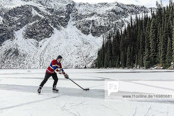 Young man playing ice hockey on lake near snow covered mountain