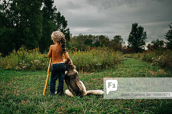 Kleiner Junge mit Waschbärfellmütze steht mit Hund auf einem Feld