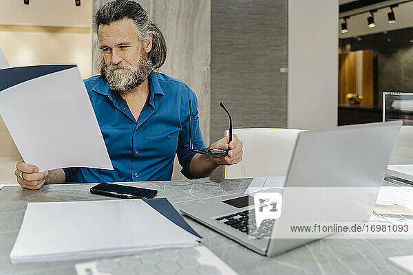 Bärtiger reifer Mann  der an seinem Schreibtisch vor seinem Laptop zu Hause einige Dokumente durchgeht. Business-Konzept