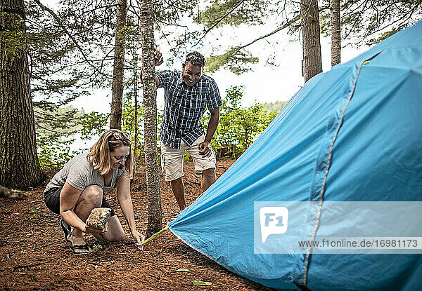 Mehrrassiges Paar lächelt und lacht beim Aufbau des blauen Zeltes  Maine