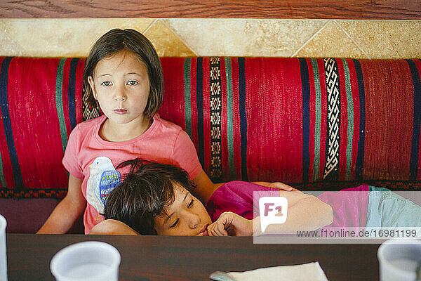 Ein kleines Mädchen sitzt an einem Restauranttisch und hat den Kopf ihres Bruders auf dem Schoß.