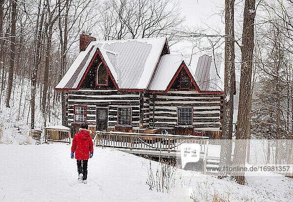 Junge in rotem Wintermantel geht an einem verschneiten Wintertag auf eine Blockhütte zu.