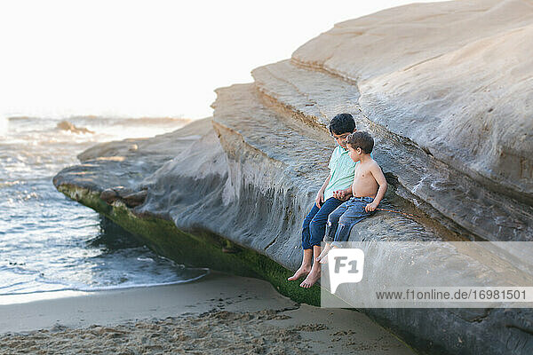 Brothers sitting on a big rock at the beach and holding hands.