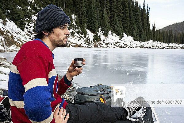 Seitenansicht eines jungen Mannes in Pullover und Mütze  der am Ufer eines zugefrorenen Sees in der Nähe eines Nadelwaldes sitzt und ein heißes Getränk trinkt  während er sich während einer Pause beim Eishockeytraining in British Columbia  C