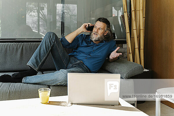 Älterer Mann mit Bart telefoniert auf einem Sofa vor seinem Laptop. Business-Konzept