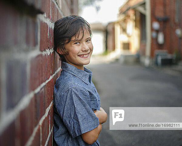 A smiling boy standing in a sunlit alley leans against brick wall