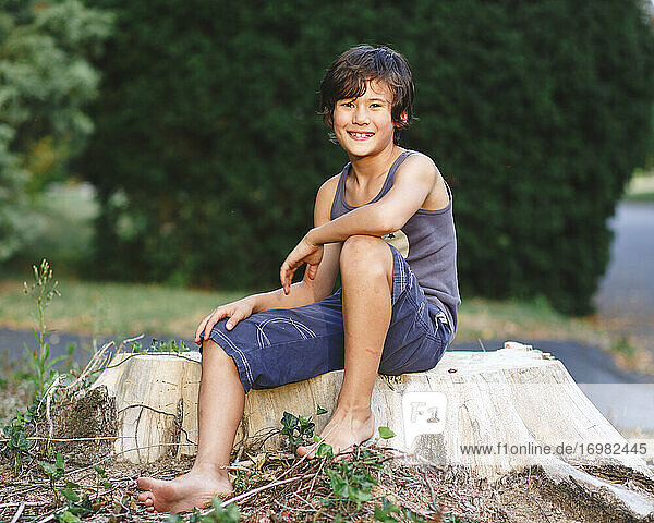 portrait of happy barefoot boy with golden skin sitting on tree stump