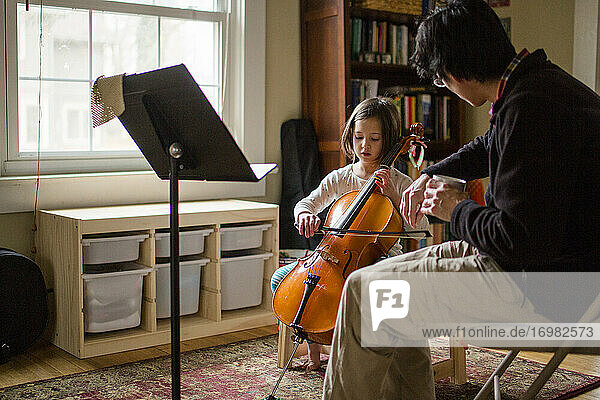 Ein Vater hilft einem kleinen Mädchen  zu Hause am Fenster Cello zu üben