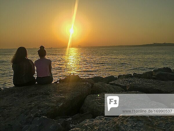 Zwei Freundinnen sitzen auf Felsen und warten auf den Sonnenuntergang an einer Küste im Sommer.