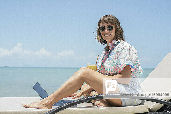 Portrait happy woman with cocktail and laptop on sunny beach lounge chair