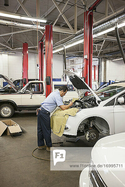 Hispanic mechanic leans on a car as he works on the engine compartment in an auto repair shop