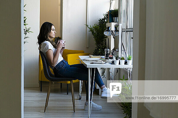 Nachdenkliche Geschäftsfrau  die eine Kaffeetasse hält und auf einem Stuhl im Büro sitzt