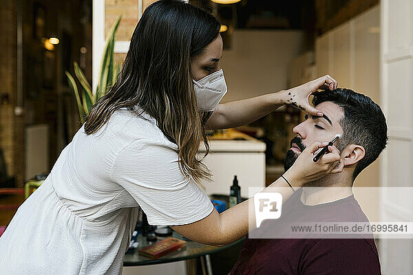 Young female stylist applying make-up to groom in salon during coronavirus