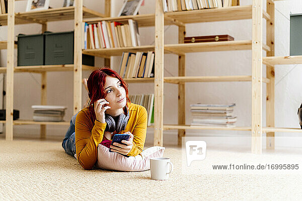 Nachdenkliche Frau mit Mobiltelefon  die wegschaut  während sie zu Hause auf dem Teppich liegt