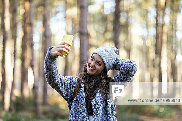 Lächelnde Frau mit Hand hinter dem Kopf  die ein Selfie macht  während sie im Wald von Cannock Chase steht