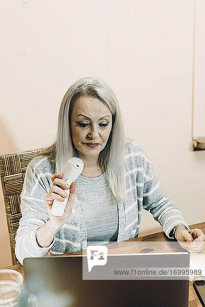 Senior woman showing infrared thermometer during online consultation at home
