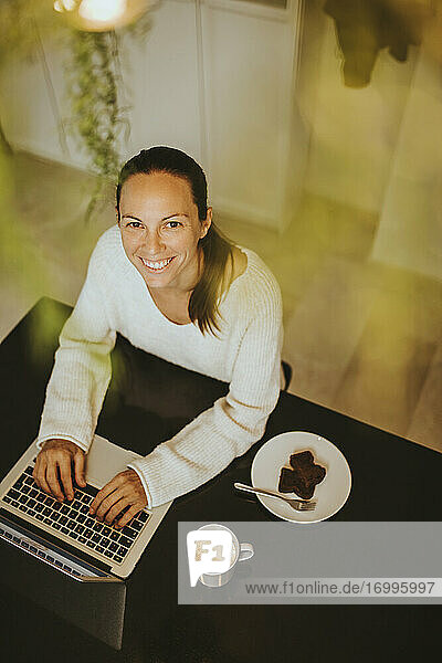 Lächelnde Frau sitzt mit Schokoladen-Brownie und Kaffee an der Kücheninsel und benutzt einen Laptop