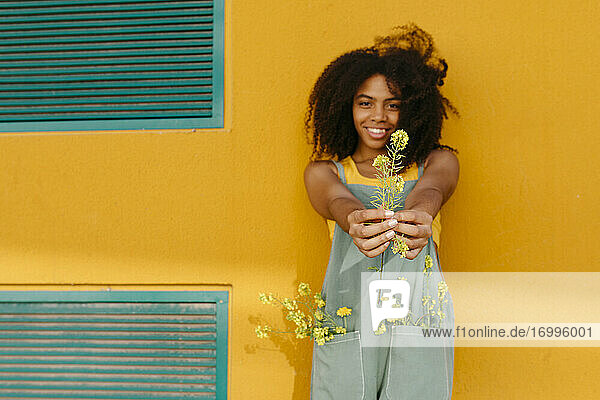 Porträt einer glücklichen jungen Frau in Latzhose mit Blumen vor einer gelben Wand