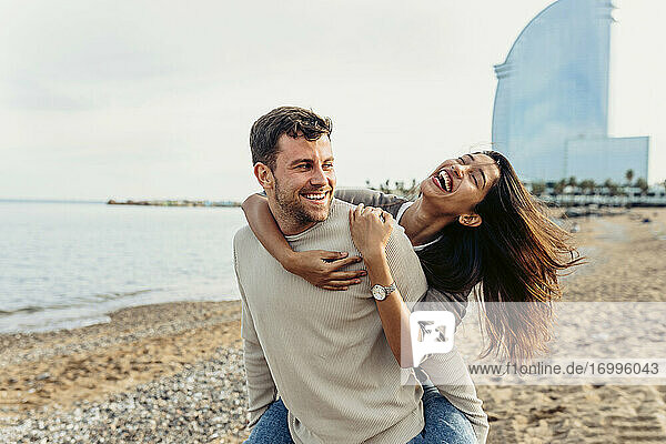 Ein lächelnder Mann nimmt eine fröhliche Frau am Strand huckepack
