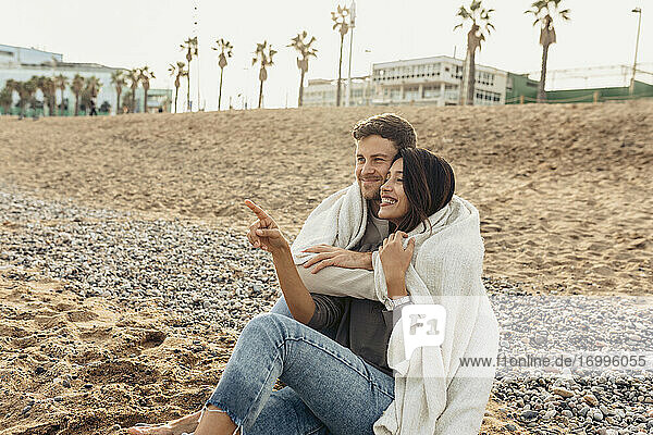Lächelnde Freundin  die auf ihren Freund zeigt  während sie ihn am Strand umarmt