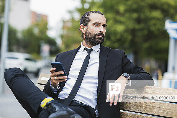 Porträt eines bärtigen Geschäftsmannes  der auf einer Bank sitzt und ein Smartphone in der Hand hält