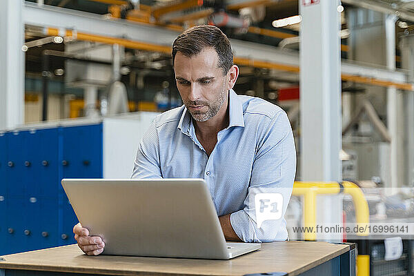Männlicher Unternehmer  der einen Laptop benutzt und sich auf einen Schreibtisch in einer Fabrik stützt