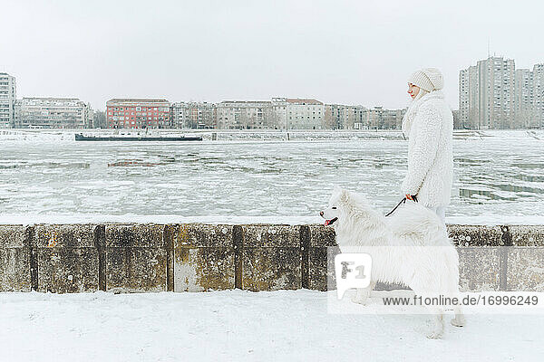 Serbia  Petrovaradin  white dressed young woman standing with white dog in the snow at riverside