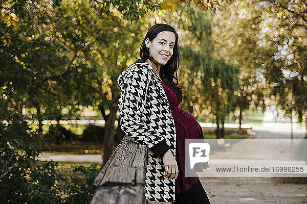 Lächelnde schwangere Frau  die sich auf eine Holzkonstruktion im Park stützt