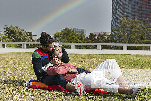 Zärtlicher Mann  der seinen Partner umarmt  während er im Park vor einem Regenbogen sitzt
