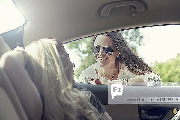Young woman leaning on car window while talking to friend during road trip