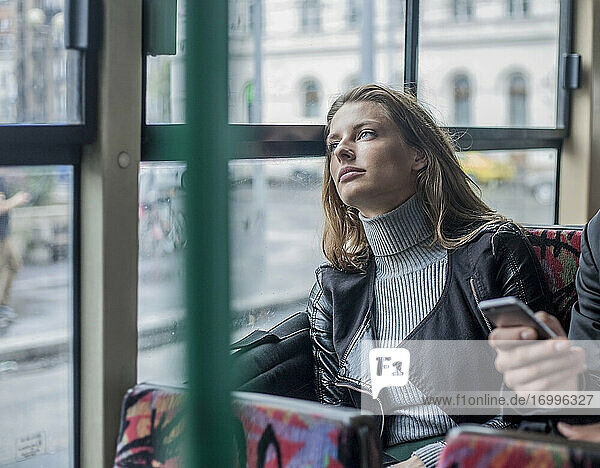 Nachdenkliche junge Geschäftsfrau  die im Bus neben einem männlichen Kollegen sitzt und sein Smartphone in der Hand hält