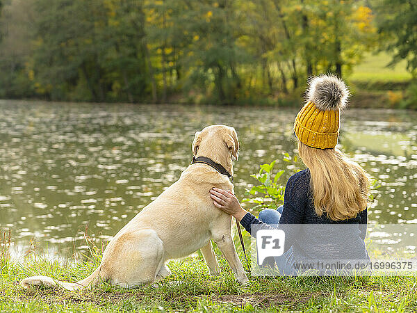 Blonde Frau sitzt mit Hund am Seeufer im Herbst