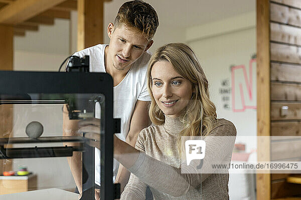 Geschäftsfrau  die zu Hause sitzend einen 3D-Drucker bedient  während ein Kollege hinter ihr steht