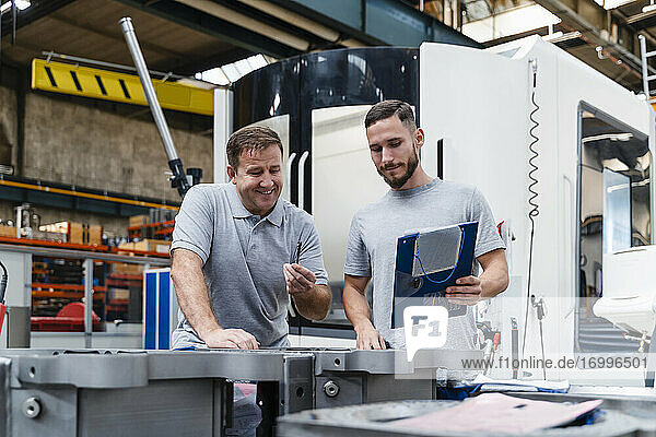 Mature male technician analyzing product standing by young colleague in industrial factory