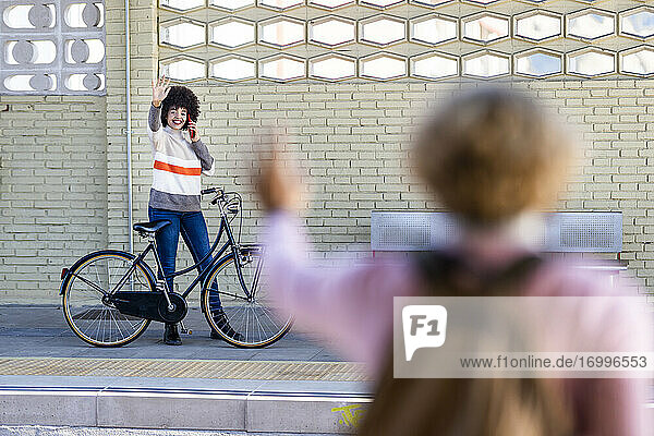 Glückliche junge Frau mit Fahrrad  die einem Freund auf dem Bahnsteig zuwinkt