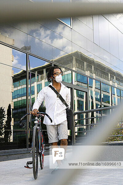 Mann mit Gesichtsmaske geht mit Fahrrad gegen modernes Gebäude während Covid-19 in der Stadt