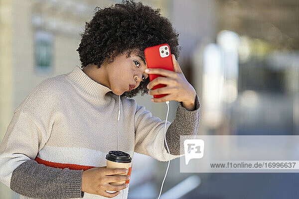 Junge Frau  die einen wiederverwendbaren Becher in der Hand hält  während sie ein Selfie mit ihrem Smartphone am Bahnhof macht