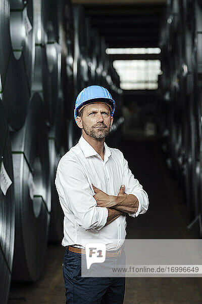 Businessman contemplating with arms crossed standing by steel rolls in factory