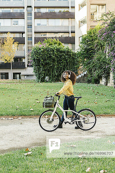 Junge Frau mit Kopfhörern  die mit dem Fahrrad auf dem Fußweg geht