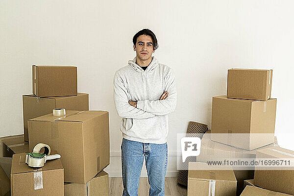 Junger Mann mit verschränkten Armen inmitten von Kartons in einem neuen Haus
