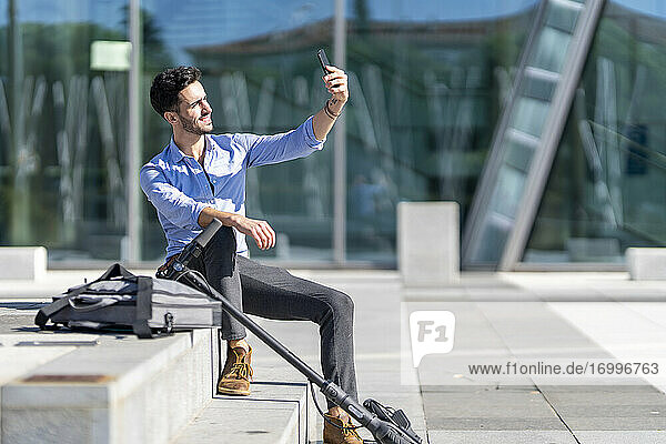 Businessman with push scooter and briefcase smiling while taking selfie sitting on steps