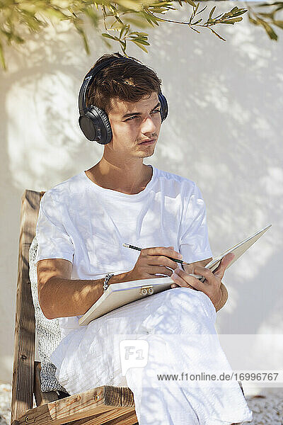Thoughtful young man with book and pen listening music through headphones against white wall