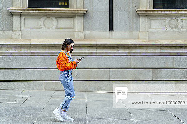 Junge Frau  die ein Mobiltelefon benutzt  während sie auf dem Gehweg gegen eine Mauer läuft