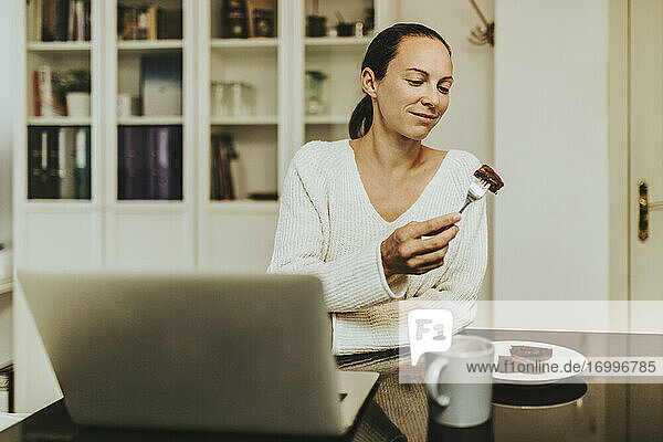 Frau isst Schokoladen-Brownie und sitzt mit Kaffee und Laptop in einer beleuchteten Küche
