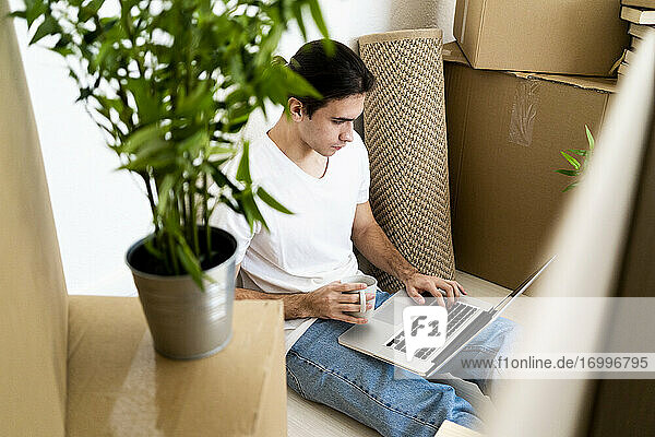 Junger Mann  der einen Laptop benutzt und eine Kaffeetasse hält  während er in seiner neuen Wohnung sitzt