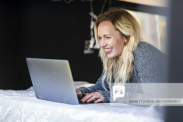 Happy woman working on laptop while lying on bed in camper van